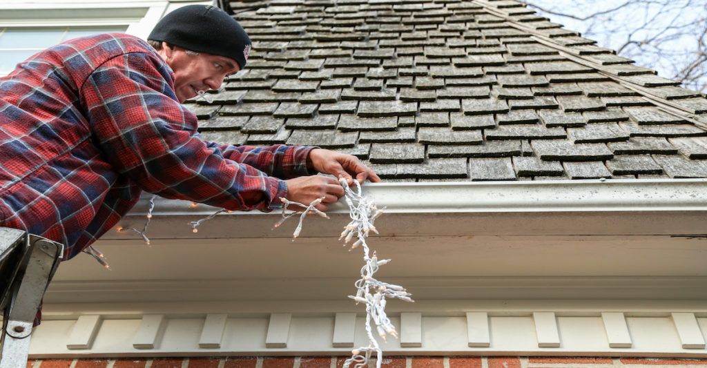 Tips for hanging holiday lights. Photo of a man on a ladder hanging Christmas lights on his house.